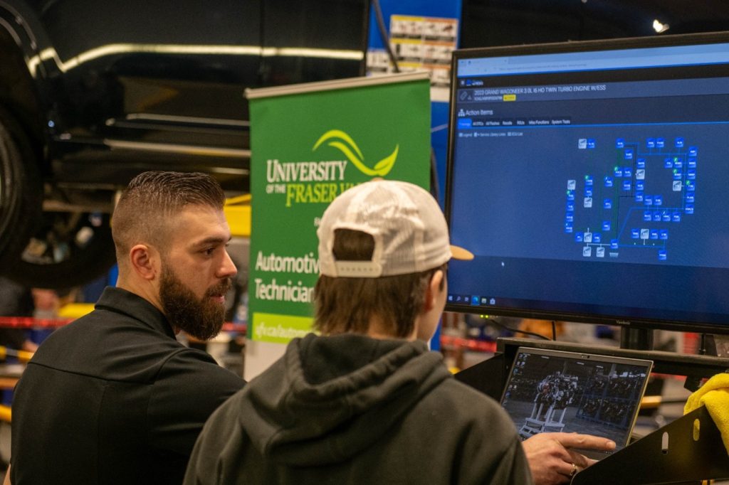 Two individuals in an automotive workshop observe a computer screen. A vertical banner for the University of the Fraser Valley Automotive Technician program is visible in the background.