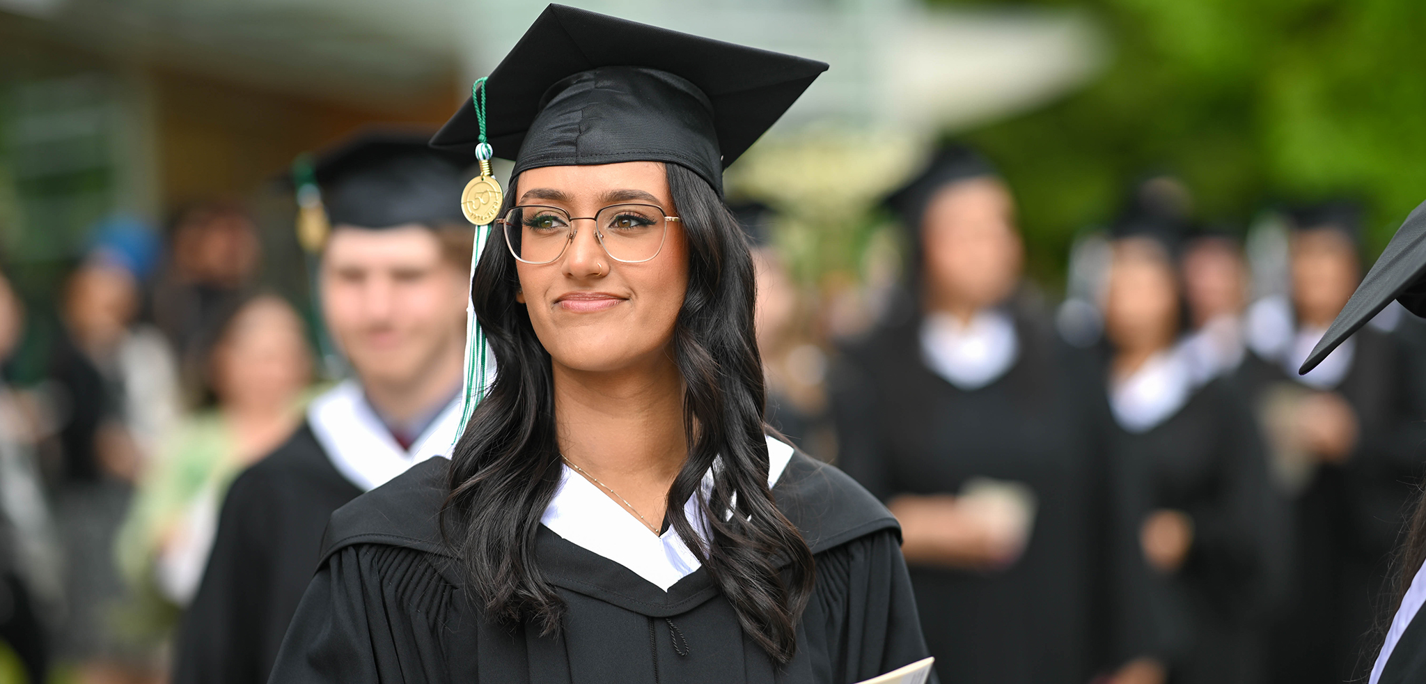 A graduate in cap and gown smiling, standing among a crowd of fellow graduates.