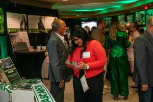 People mingle at a University of the Fraser Valley (UFV) event, surrounded by informational displays and green lighting. A woman in a red jacket and name tag looks at a pamphlet near a table decorated with UFV Cascades materials, while a man in a gray suit stands beside her. Other attendees chat in the background, some dressed in formal and colorful attire. The atmosphere is warm and social.