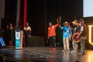 A group of people stand on stage during a ceremony. Four Indigenous men are positioned on the right, wearing traditional hats and holding hand drums and paddles, appearing to perform or sing. To the left, a man stands at a podium with the University of the Fraser Valley (UFV) logo.