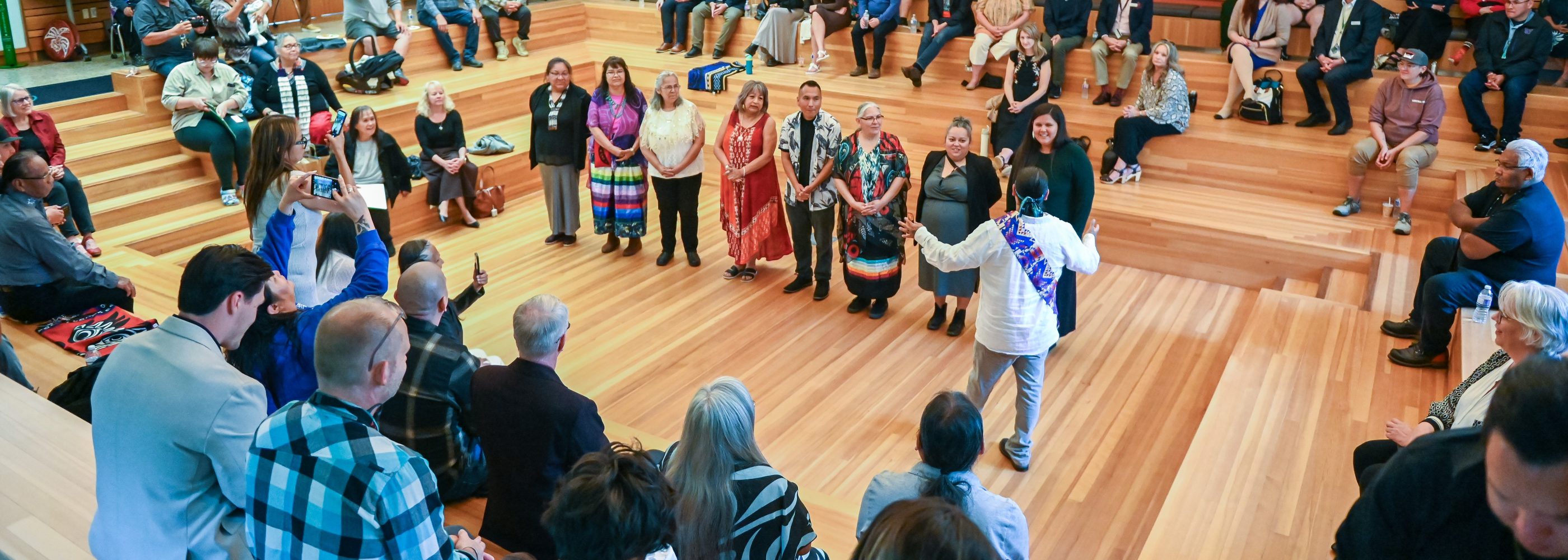 A group of people gathered in a circular wooden amphitheatre during an Indigenous cultural event, with several individuals standing at the center in traditional attire while others watch and take photos from the surrounding benches.