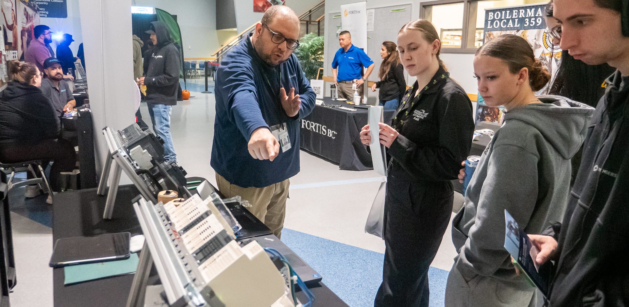 A man gestures and points while explaining technical equipment displayed on a table to a small group of students at an indoor career or industry event. Booths, banners, and other attendees are visible in the background, creating a hands on learning and networking environment.