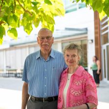 An older couple stands outdoors on the UFV campus, posing together in front of a modern building. The man wears glasses and a blue short sleeve button up shirt, and the woman wears a pink patterned jacket over a light top. Green tree leaves frame the top of the image, and a student walks in the background near the building entrance.