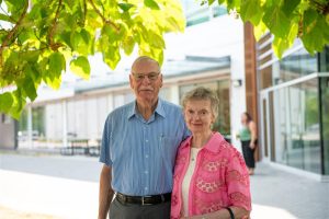 An older couple stands outdoors on the UFV campus, posing together in front of a modern building. The man wears glasses and a blue short sleeve button up shirt, and the woman wears a pink patterned jacket over a light top. Green tree leaves frame the top of the image, and a student walks in the background near the building entrance.