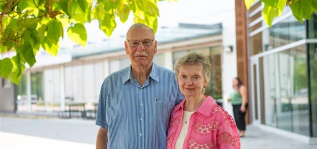 An older couple stands outdoors on the UFV campus, posing together in front of a modern building. The man wears glasses and a blue short sleeve button up shirt, and the woman wears a pink patterned jacket over a light top. Green tree leaves frame the top of the image, and a student walks in the background near the building entrance.