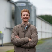 Portrait of a man in a brown jacket standing confidently on a gravel path beside farm silos and agricultural buildings.
