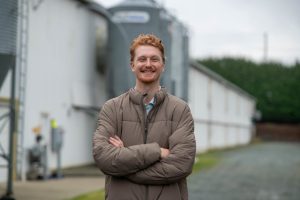Portrait of a man in a brown jacket standing confidently on a gravel path beside farm silos and agricultural buildings.