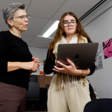 Two women standing in an office classroom, looking at a laptop together, with computers and student artwork visible in the background.