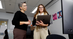Two women standing in an office classroom, looking at a laptop together, with computers and student artwork visible in the background.