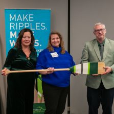 Three people stand indoors holding a large decorative paddle during a presentation or recognition event. Behind them is a teal banner that reads “Make Ripples. Make Waves.” with University of the Fraser Valley branding. Two additional paddles are displayed upright on a stand in the background.