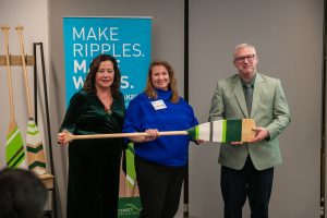 Three people stand indoors holding a large decorative paddle during a presentation or recognition event. Behind them is a teal banner that reads “Make Ripples. Make Waves.” with University of the Fraser Valley branding. Two additional paddles are displayed upright on a stand in the background.