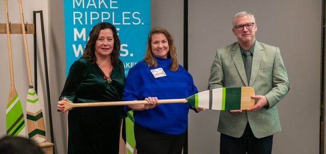 Three people stand indoors holding a large decorative paddle during a presentation or recognition event. Behind them is a teal banner that reads “Make Ripples. Make Waves.” with University of the Fraser Valley branding. Two additional paddles are displayed upright on a stand in the background.