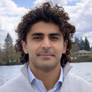 Man with dark hair is standing outdoors in front of a calm body of water, with trees and a cloudy sky in the background. They are wearing a light-colored jacket over a collared shirt and looking directly at the camera with a neutral expression. Only head and top of shoulders are visible.