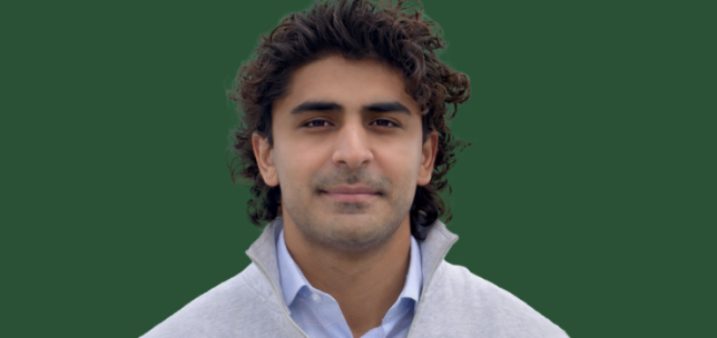 Man with dark hair is in front of a dark green background. They are wearing a light-gray colored jacket over a collared shirt and looking directly at the camera with a neutral expression. Only head and top of shoulders are visible.