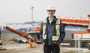 A smiling man stands at an active industrial construction site wearing a white hard hat, navy jacket, and black high-visibility safety vest with bright yellow reflective stripes. Behind him, large red-and-white material processing and conveyor equipment stretches across the site, surrounded by gravel piles, construction vehicles, concrete blocks, and electrical equipment. Dark, forested mountains rise in the distance, partially obscured by low clouds and mist beneath an overcast sky.