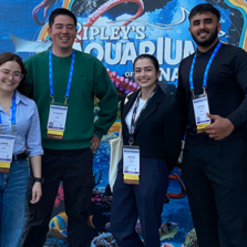 Four conference attendees wearing event badges stand together outdoors, smiling at the camera in front of a large, colorful Ripley’s Aquarium of Canada display. The ocean-themed backdrop features an octopus, jellyfish, fish, coral, and other marine life in vivid shades of blue and purple. The group is dressed in a mix of business-casual and professional attire, with an urban plaza and buildings visible in the background.