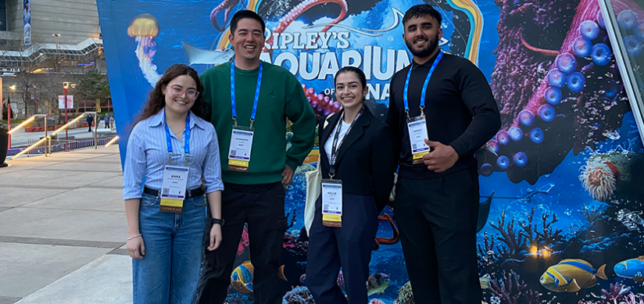 Four conference attendees wearing event badges stand together outdoors, smiling at the camera in front of a large, colorful Ripley’s Aquarium of Canada display. The ocean-themed backdrop features an octopus, jellyfish, fish, coral, and other marine life in vivid shades of blue and purple. The group is dressed in a mix of business-casual and professional attire, with an urban plaza and buildings visible in the background.