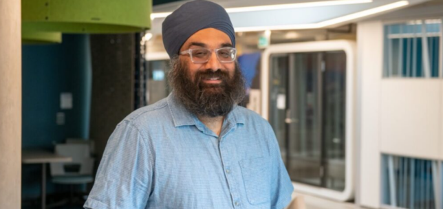 A smiling man wearing a dark blue turban, glasses, and a light blue short-sleeved button-up shirt stands in a bright, modern office interior. He has a full beard and is positioned in front of contemporary architectural features, including curved ceiling details, green and dark teal accents, glass-walled rooms, and an open hallway.