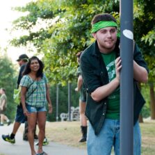 A student wearing a green bandana holds onto a light pole during an outdoor campus activity, while several other students stand and walk nearby on a tree-lined path.