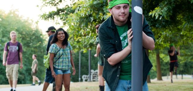 A student wearing a green bandana holds onto a light pole during an outdoor campus activity, while several other students stand and walk nearby on a tree-lined path.