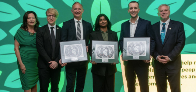 Six individuals stand together on stage in front of a bright green backdrop. Three hold framed awards featuring Indigenous artwork, while the group smiles for a photo.