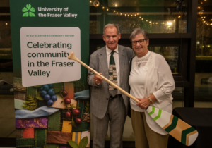 Two individuals smile while holding a paddle featuring green and white designs, standing beside a UFV banner promoting the St’elt’elawtexw Community Report, Celebrating Community in the Fraser Valley.