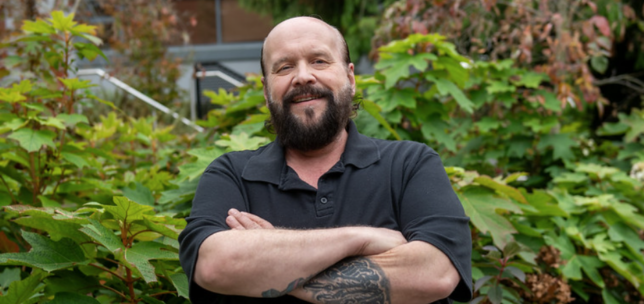 A smiling person with a beard stands outdoors with their arms crossed, wearing a black polo shirt and surrounded by lush greenery.